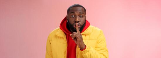 Waist-up serious-looking african-american bearded man in yellow jacket red hoodie bend camera asking keep quiet say shhh show shush gesture index finger on lips, confidential information taboo photo