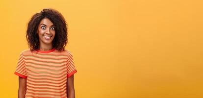 Waist-up shot of amazed and excited charming african american woman with curly hairstyle popping eyes from thrill and joy smiling broadly being surprised by great gift over orange background photo