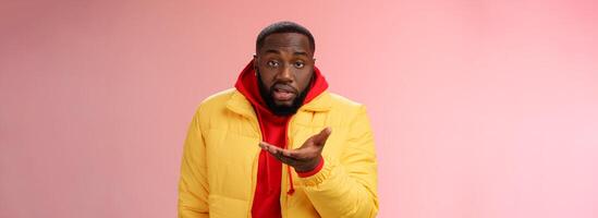 Bothered rude ignorant african-american bearded man pointing palm camera look dumb perplexed, standing confused cannot get clue what happening, wearing yellow jacket, pink background photo