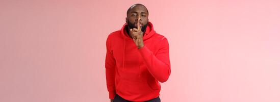Shhh it taboo. Portrait sensual funny cute african-american bearded man in red hoodie bending camera show shush gesture index finger on lips telling secret asking keep quiet not tell anyone mystery photo