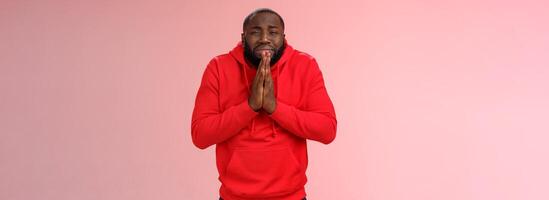 Miserable african american man beard in red hoodie stooping make supplicating upset grimace grying sobbing asking help say please hold hands pray begging mercy, apologizing need money photo