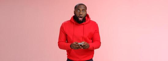 Amused overhwelmed black bearded guy in red hoodie drop jaw gasping widen eyes staring camera impressed holding smartphone using amazing cool game astonished new phone features, standing pink wall photo