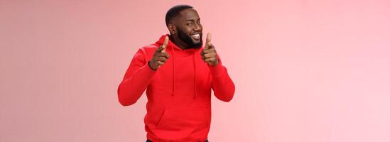 Nice work well done friend. Proud cheering good-looking cheeky young flirty black guy beard in red hoodie pointing finger pistols camera smug congratulating mate good game, standing pink background photo