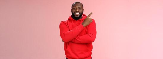 Cheeky outgoing smiling attractive african-american male model in red hoodie grinning energized pointing upper left corner indicating awesome product recommend promo, standing pink background photo