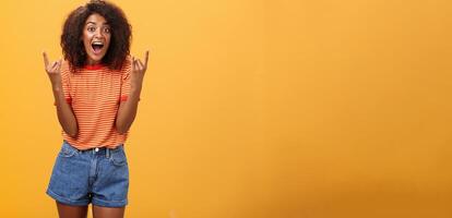 Stylish african american woman being delighted and excited before cool concert showing rock n roll gesture with both hands screaming from satisfaction and happiness cheering over orange background photo