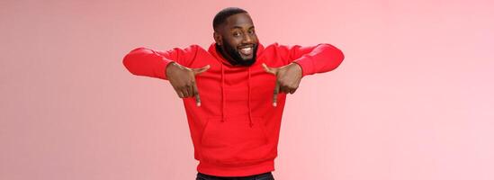 Sassy young charismatic handsome bearded black guy in red hoodie pointing down index fingers smiling broadly showing awesome copy space solution cool product for you, standing pink background photo