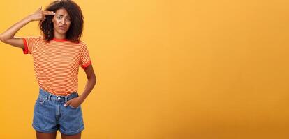 Portrait of displeased gloomy and sad annoyed dark-skinned female with curly hairstyle showing gun with hand near temple frowning wanting to kill herself from exhaust and boredom over orange wall photo