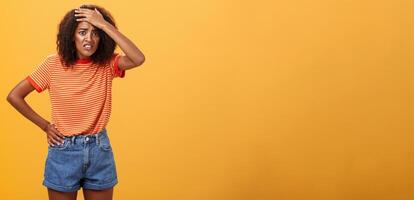 mierda yo a.m en problema. preocupado ansioso linda de piel oscura mujer en de moda camiseta y pantalones cortos apretando dientes con culpable Mira ceñudo puñetazos frente teniendo problema o problema terminado naranja pared foto