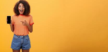 Impressed fascinated stylish slim african american curly-haired woman in striped t-shirt dropping jaw from amazement holding awesome smartphone pointing at device screen over orange wall photo