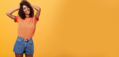 Girl feeling awesome making cool hairstyle posing near mirror feeling glad to have date tonight holding hands on curly hair smiling joyfully and tender standing against orange background photo