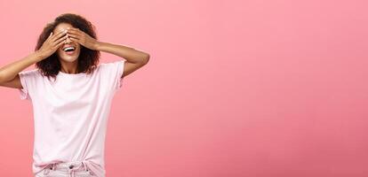 Portrait of charming joyful dark-skinned playful woman with curly hair in t-shirt closing eyes and counting ten with broad happy smile playing hide n seek or waiting for surprise over pink background photo