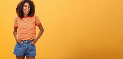 Woman sometimes wants feel childish. Joyful enthusiastic and charismatic young african american female with afro hairstyle winking happily showing tongue holding hands in pockets against orange wall photo