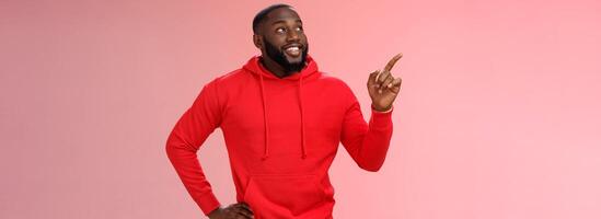 Happy charismatic delighted african-american bearded guy smiling white teeth look pointing intrigued upper left corner curious what interesting product standing impressed pink background photo