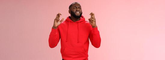 Passionate devoted african-american male fan praying cross fingers good luck asking god win bet biting lip impatiently waiting important results supplicating hopefully, standing pink background photo