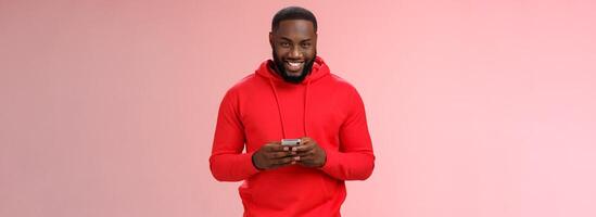 Attractive happy smiling african american bearded guy in red hoodie playing games smartphone grinning joyfully messaging having interesting online chat, standing satisfied pink background photo