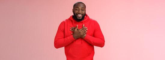 Pleased touched african american smiling young bearded man hold hands heart grinning thanking heartwarming lovely congratulations, receive good feedback standing happy thanking everyone photo