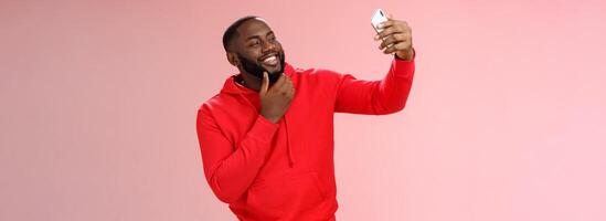 Cheeky cool african american confident bearded guy in red hoodie taking selfie update date app profile photo holding smartphone raised touching face sassy smiling look phone display flirty