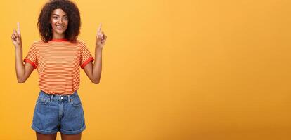 Looking only up and forward. Optimistic ambitious stylish dark-skinned female student in striped cool t-shirt and shorts raising hands pointing upwards and smiling friendly over orange wall photo
