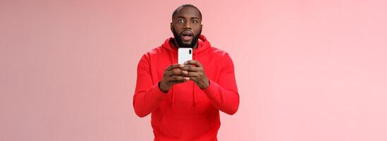 Impressed focused handsome african american bearded guy looking intrigued holding smartphone recording video capturing awesome fight, widen eyes curious surprised, standing pink background photo