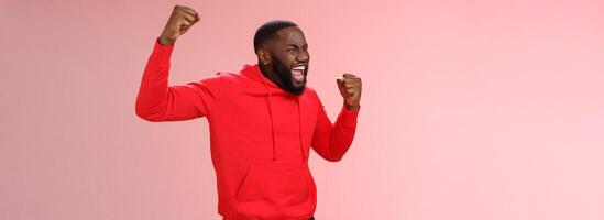 Extremely happy bearded young 25s black guy triumphing happily yelling yeah raising clenched fists celebrating success winning bet lottery standing happily pink background cheer receive great news photo
