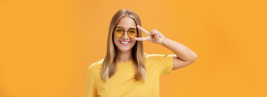 Cute optimistic and friendly young caucasian woman with fair hair in yellow t-shirt and sunglasses showing peace sign near eye and smiling cheerfully at camera taking shot over orange background photo