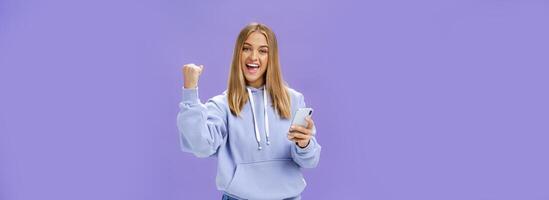 Yes we did it. Pleased excited woman in hoodie raising fist in celebration and success gesture smiling broadly triumphing holding smartphone reacting to positive news in cellphone over purple wall photo