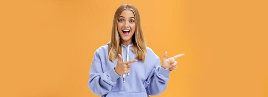 Amused girl pointing at curious copy space showing left with index fingers smiling excited and surprised standing entertained with upbeat grin in cozy blue hoodie over orange background photo