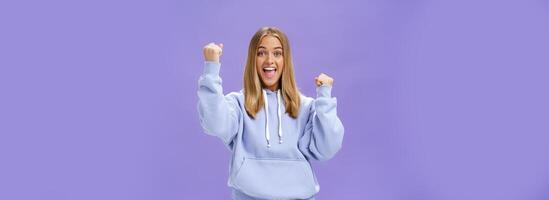 Cheerful happy and supportive young girlfriend with fair hair and tan in warm hoodide raising fists in cheer and triumph smiling saying yeah celebrating goal or success over purple background photo