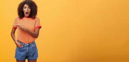 Studio shot of offended shocked stylish woman being robbed fasping and staring nervously begging stranger help out pointing left concerned and worried frowning, posing displeased over orange wall photo