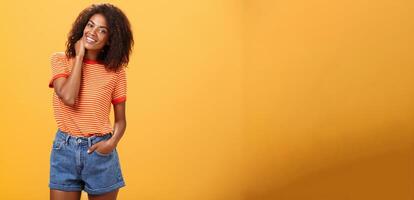 Stylish African American slim woman with curly medium hair holding hand in denim shorts tilting head, rubbing neck while smiling friendly feeling uncomfortable or shy in new company over orange wall photo