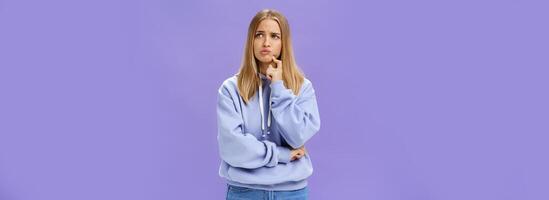 Silly concerned girlfriend trying think up how solve problem standing thoughtful with troubled expression touching jaw pouting, frowning staring right, thinking making up decision over purple wall photo