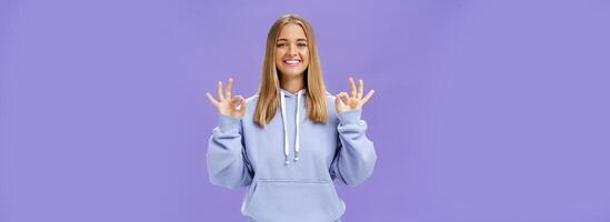 Girl got everything under control. Delighted happy charming woman with fair hair and tanned skin without makeup showing okay gesture smiling assured and pleased posing in hoodie over purple wall photo