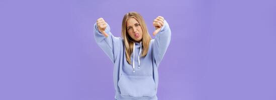 Indoor shot of moodie displeased and unimpressed cool modern female with blond hair and tanned skin tilting head sticking out tongue showing thumbs down in dissatisfaction posing against purple wall photo