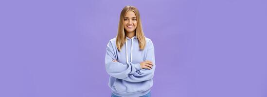 Portrait of charming charismatic european woman with tanned skin in trendy over-sized hoodie crossing hands against chest smiling broadly at camera standing self-assured against purple background photo