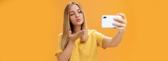 elegante glamoroso hembra Moda blogger finalizando grabación vídeo vía teléfono inteligente por enviando aire Beso a cámara, tomando selfie con sensual y confidente mirada a pantalla posando terminado naranja pared foto