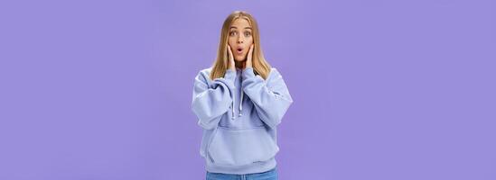 Indoor shot of amused and thrilled surprised good-looking young woman in stylish hoodie folding lips in wow sound popping eyes at camera shocked and excited learning amazing news over purple wall photo