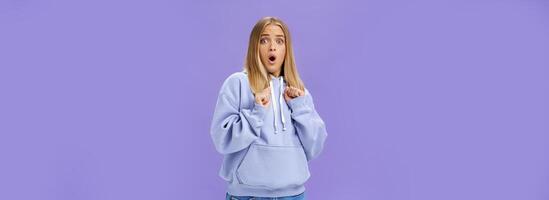 Woman scared of sudden loud noise in dark gasping looking shocked and concerned tilting backwards with frightened look pressing clenched fists to body from fear, wearing warm hoodie over purple wall photo