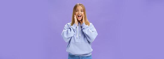 Woman joyfully smiling at camera and touching cheeks as if being surprised and happy see friend meeting person in airport feeling cheerful to reunite standing over purple background in cute hoodie photo
