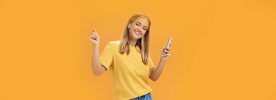 Joyful carefree european woman wearing wireless earbuds holding smartphone dancing with raised hands and pleased happy smile leading vanlife having time to relax and enjoy life over orange wall photo