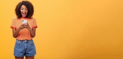 Girl expressing excitement and joy receiving awesome invitation via messages yelling from delight and happiness looking at smartphone screen impressed and thrilled over orange background photo