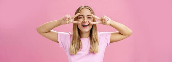 Joyful friendly and peaceful common european woman with blond hair in casual t-shirt showing peace or disco signs over eyes smiling cheerfully at camera having fun against pink background photo