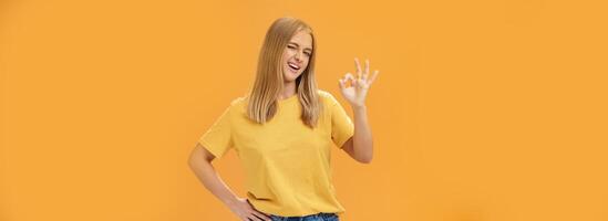 Confident and skillful female student assuring teacher she do great job holding hand on waist winking at camera with self-assured look and showing okay gesture assuring everything ok over orange wall photo