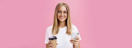 Pleasant friendly-looking girl smiling at camera holding paper cup of coffee and smartphone. Portrait of joyful nice woman drinking morning drink, posing opinion about cafe in internet over pink wall photo