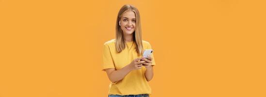 Young caucasian girl with tanned skin and fair hair using wireless earphones to call friend via smartphone holding cellphone against chest, smiling cheerfully at camera getting used to new technology photo