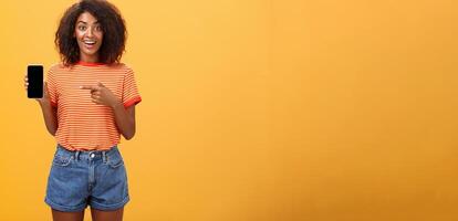 Cute african american stylish woman showing friend new app in smartphone pointing at cellphone screen with index finger smiling thrilled and happy delighted with cool device purchase over orange wall photo