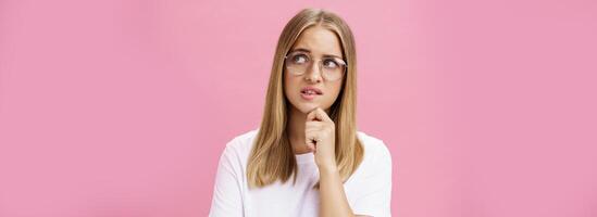 Studio shot of insecure smart nerdy woman in glasses and white t-shirt standing troubled and worried touching chin looking bothered at upper right corner hesitating, thinking against pink wall photo