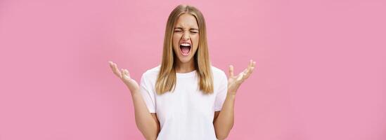 Woman feeling fed up having mental breakdown during deadline yelling with closed eyes, raising hands aside in pissed gesture feeling distressed and pressured losing temper over pink wall photo