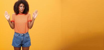 Silly insecure and sad dark-skinned female model in trendy striped t-shirt and shorts raising arms in surrender frowning being uninvolved and unaware standing clueless over orange background photo