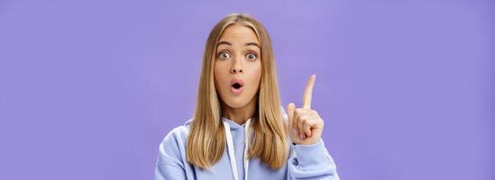 Oh I know idea. Excited cute young woman with tan in hoodie raising index finger in eureka gesture folding opened lips and staring amazed at camera having suggestion what do over blue background photo