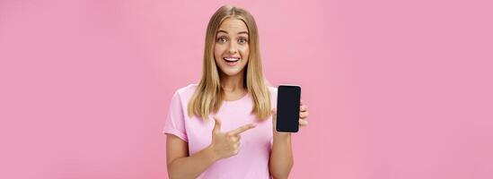 alegre atractivo y agradable mujer promoviendo frio aplicación o teléfono inteligente participación Teléfono móvil y señalando a dispositivo pantalla sonriente divertido y impresionado en pie terminado rosado antecedentes foto